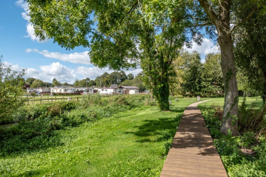 Ringwood | Direct Cremations Wooden path along greenery, with trees and a distant field under a blue sky.